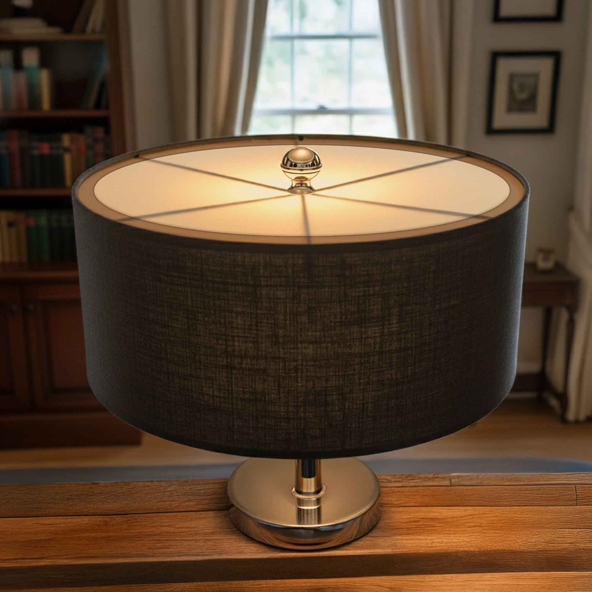 A table lamp with a dark gray textured fabric drum shade, a polished silver metal base, and a warm glowing light diffuser, set on a wooden table in a cozy, softly lit room.