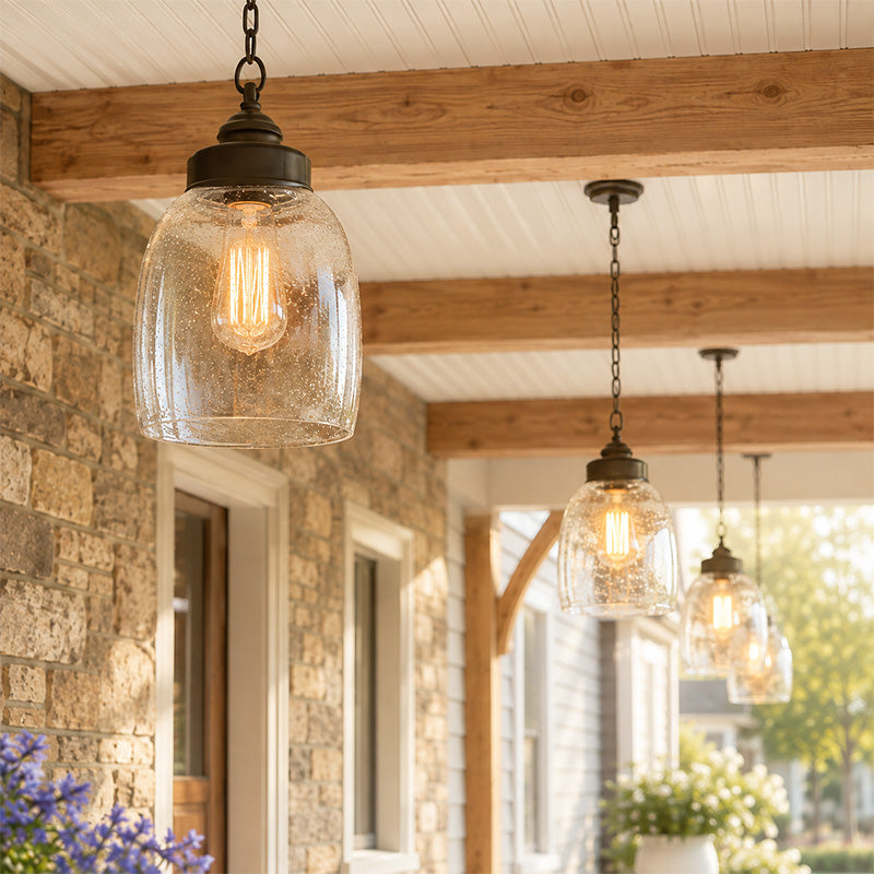 Three outdoor pendant light fixtures with clear seeded glass shades and dark metal hardware hang over a wooden-beamed porch with stone walls.